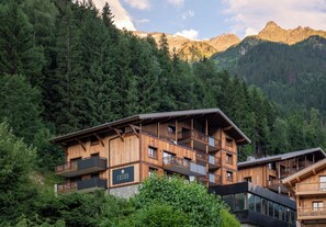 A multi-story wooden chalet with balconies, set against a backdrop of dense forest and mountains. - MGM Hôtels et Résidences - Hotel Laska (Les Contamines-Montjoie)