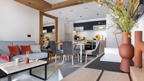 A modern living room with a sofa, coffee table, and chairs. A kitchen is visible in the background with cabinets and appliances. - MGM Hôtels et Résidences - Hotel Laska (Les Contamines-Montjoie)