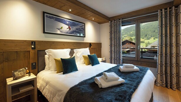A hotel room with a large bed, wooden headboard, a painting of a skier, and a window with curtains. - MGM Hôtels et Résidences - Hotel Laska (Les Contamines-Montjoie)