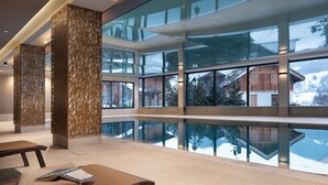 A modern indoor swimming pool with clear glass walls, a wooden floor, and a seating area with a bench and towels. - MGM Hôtels et Résidences - Hotel Laska (Les Contamines-Montjoie)