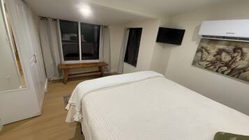 A bedroom with a bed, a bench, a TV, and an air conditioner.