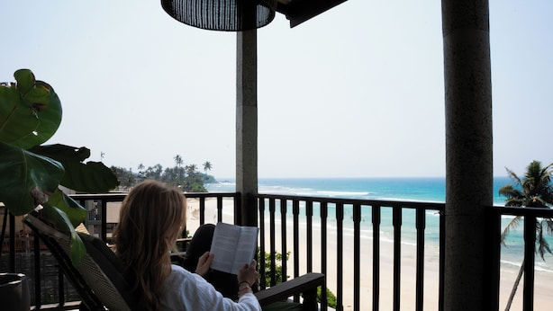 Eine Person liest auf einem Balkon mit Blick auf den Strand und das Meer.