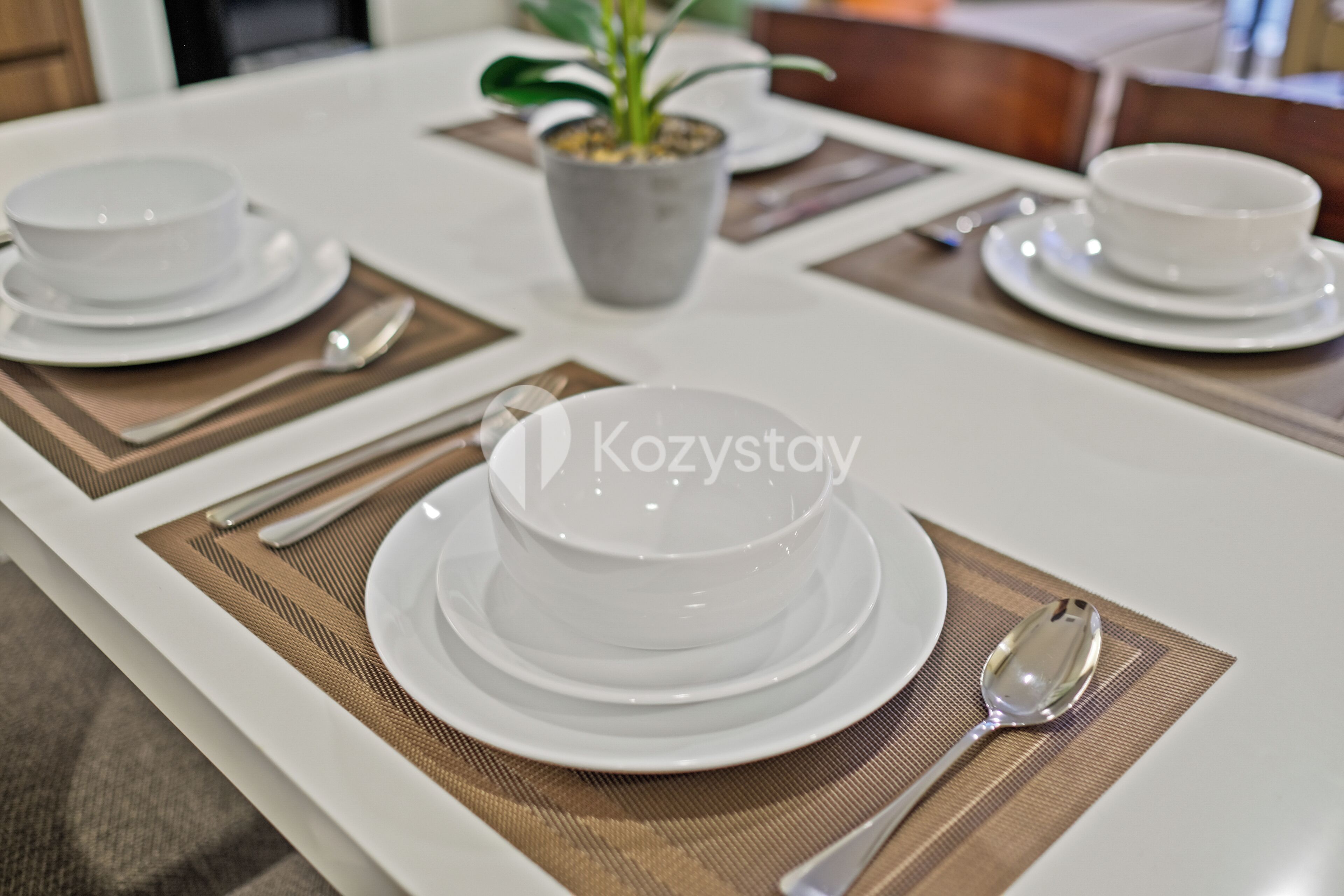Uma mesa de jantar posta com pratos, tigelas e talheres brancos, com um vaso de planta ao fundo.