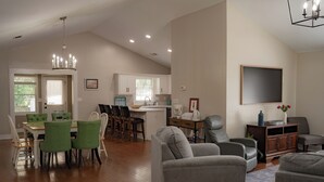 Un salón amplio con una mesa de comedor, sillas verdes, un sofá gris y un televisor montado en la pared.