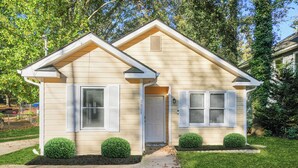 A two-story house with a beige exterior, white shutters, and a small front yard with trimmed bushes. - The ATL Escape (Atlanta)