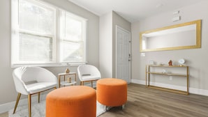 A modern living room with a large window, two white chairs, an orange ottoman, a gold-framed mirror, and a wooden console table. - The ATL Escape (Atlanta)