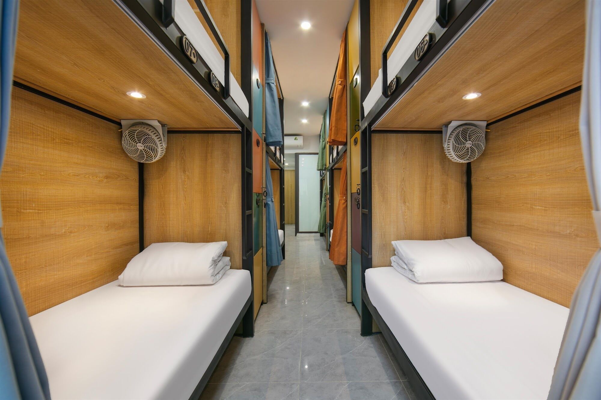 A narrow corridor with two beds on either side, each with a fan and a small window.