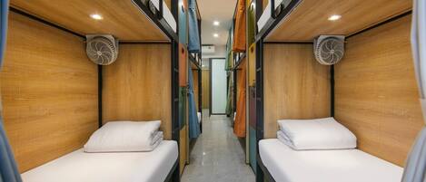 A narrow corridor with two beds on either side, each with a fan and a small window.