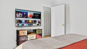 Quarto com televisão exibindo interface de serviço de streaming, mesa de cabeceira com gavetas, cama com manta vermelha e porta branca com venezianas.