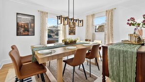 Uma área de refeições com uma mesa de madeira posta para quatro pessoas, rodeada por cadeiras castanhas. Uma luminária pendente com lâmpadas aparentes está sobre a mesa.