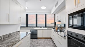A modern kitchen with a built-in oven, a dishwasher, and a large window offering a view of the ocean at sunset. - Luxury Oceanview Penthouse at Northshore Tower Cairns (Cairns North)