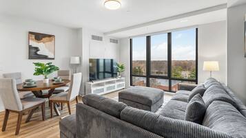A modern living room with a large grey sofa, a dining table with chairs, a TV, and a large window offering a view of trees and buildings.