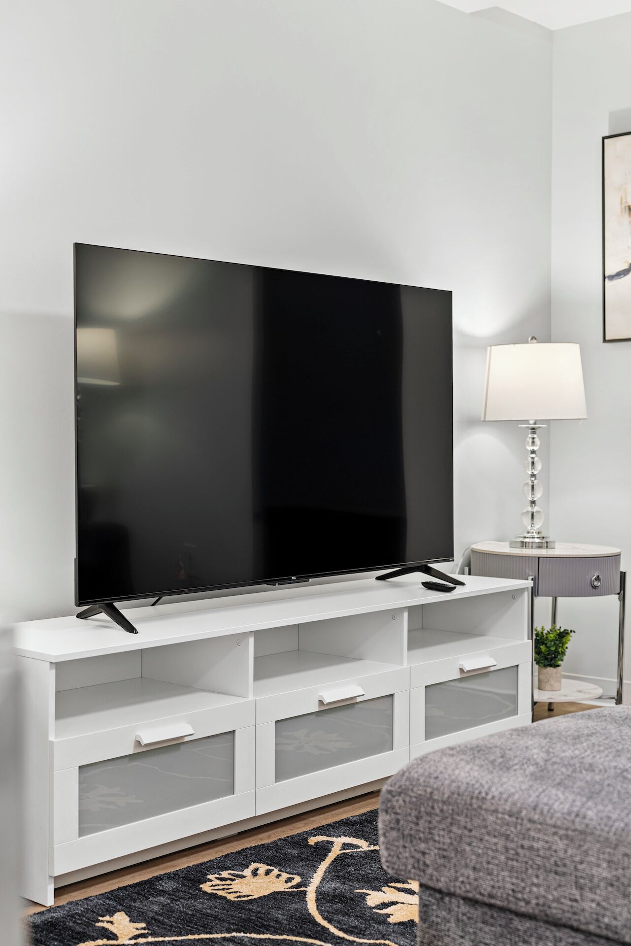 Moderni olohuone, jossa on suuri, seinälle asennettu litteänäyttöinen televisio, valkoinen TV-taso, pieni sivupöytä lampun kanssa ja harmaa sohva.