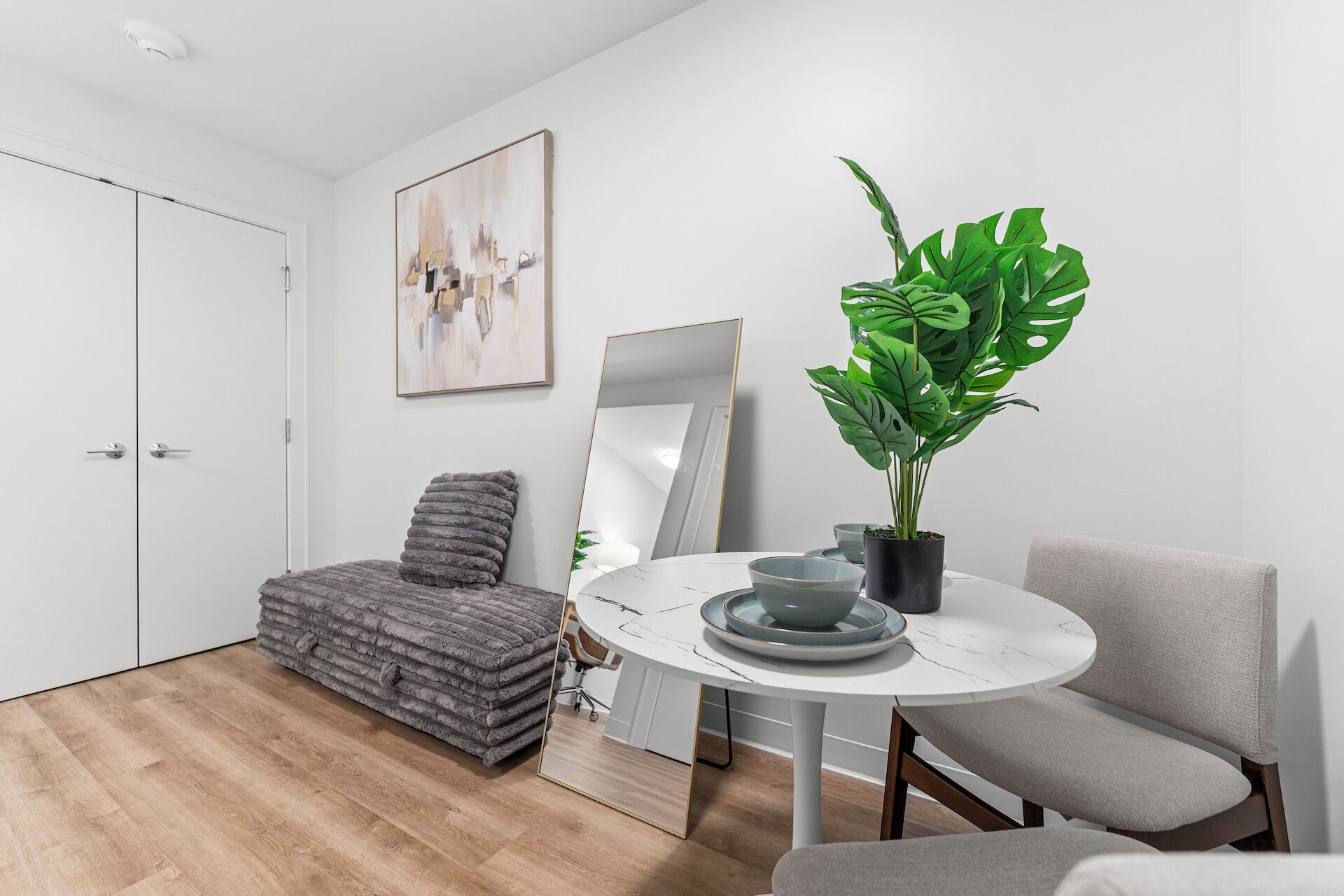 Ein modernes Wohnzimmer mit einem runden Esstisch, grau gepolsterten Sesseln, einem grauen Sofa, einer großen Kühlpflanze, einem Spiegel und einem Gemälde an der Wand.