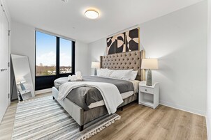 A modern bedroom with a large bed, two bedside tables, a view through the window, and a cityscape outside.