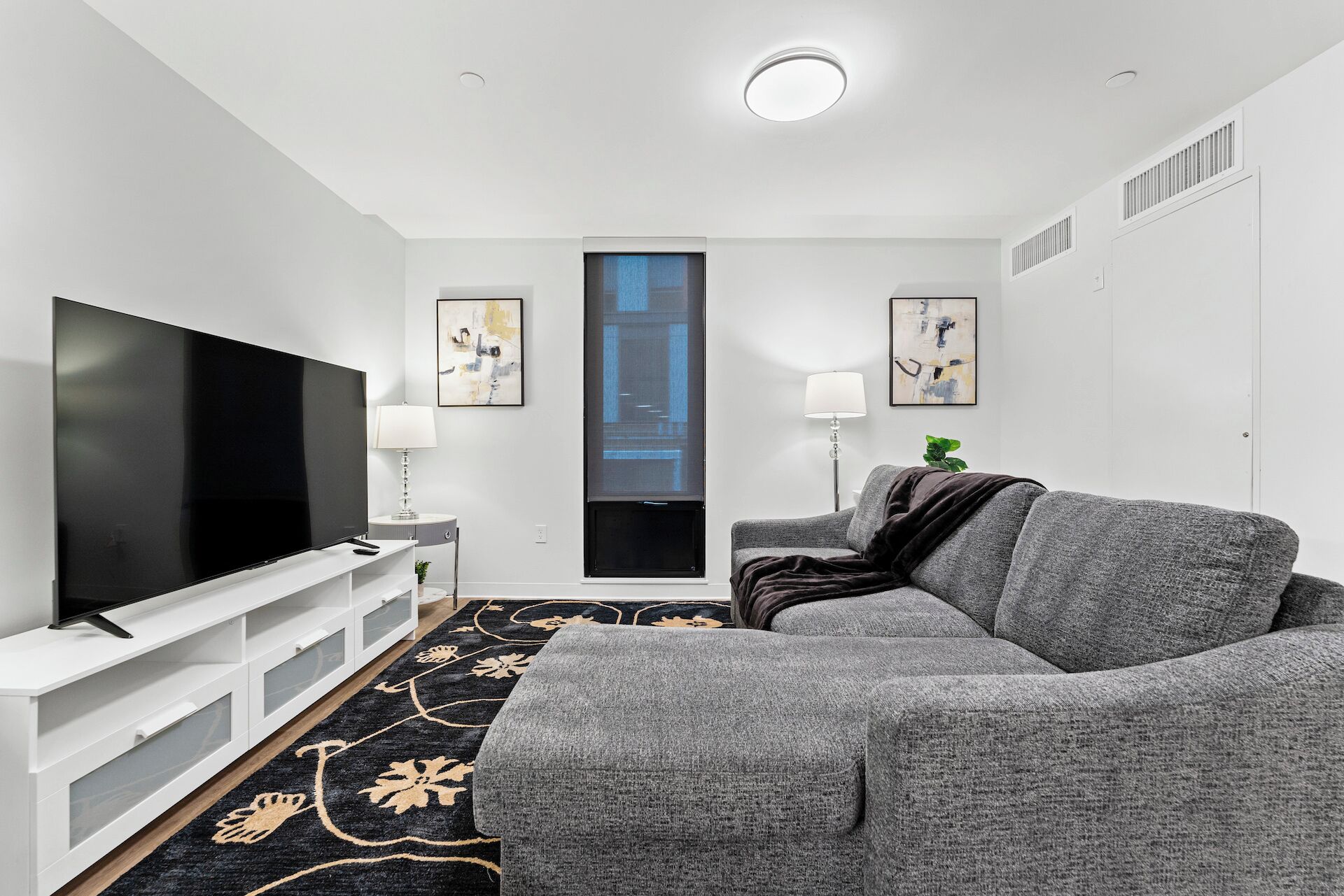 Una sala de estar moderna con televisor de pantalla plana, sofá gris, mueble blanco para entretenimiento y alfombra estampada en negro y dorado.