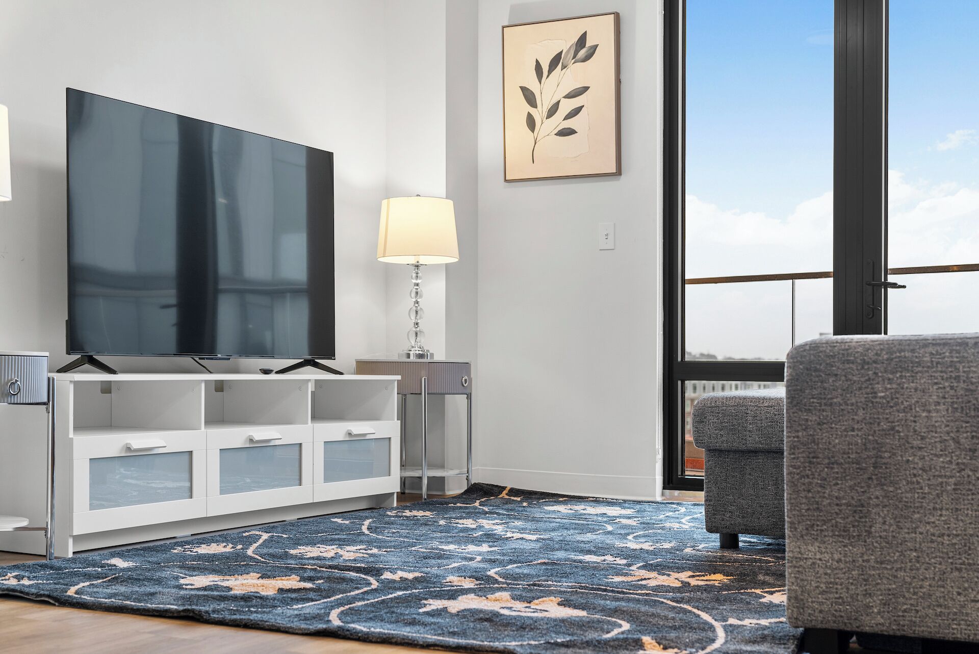 A modern living room with a flat-screen TV, a white cabinet, a lamp, a gray armchair, and a large window with a view.