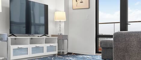 A modern living room with a flat-screen TV, a white cabinet, a lamp, a gray armchair, and a large window with a view.