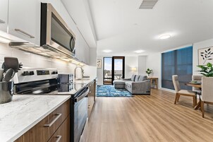 A modern kitchen with a built-in oven, microwave, and toaster. There is a dining area with a sofa and chairs, and a large window offering a view of the city.