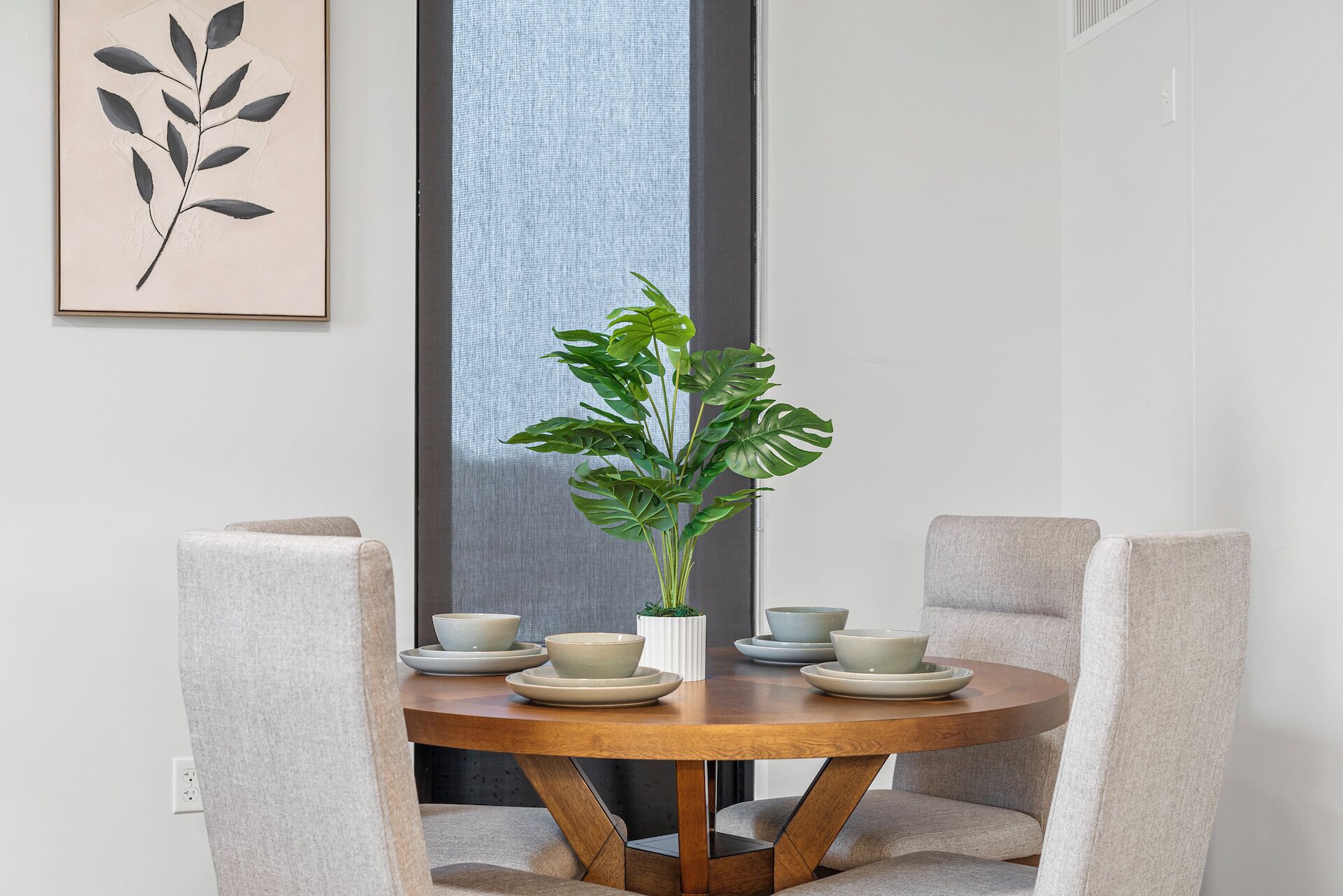 Une table de salle à manger ronde en bois, entourée de quatre chaises grises à revêtement rembourré, un pot contenant une plante monstera et quatre bols posés sur la table.
