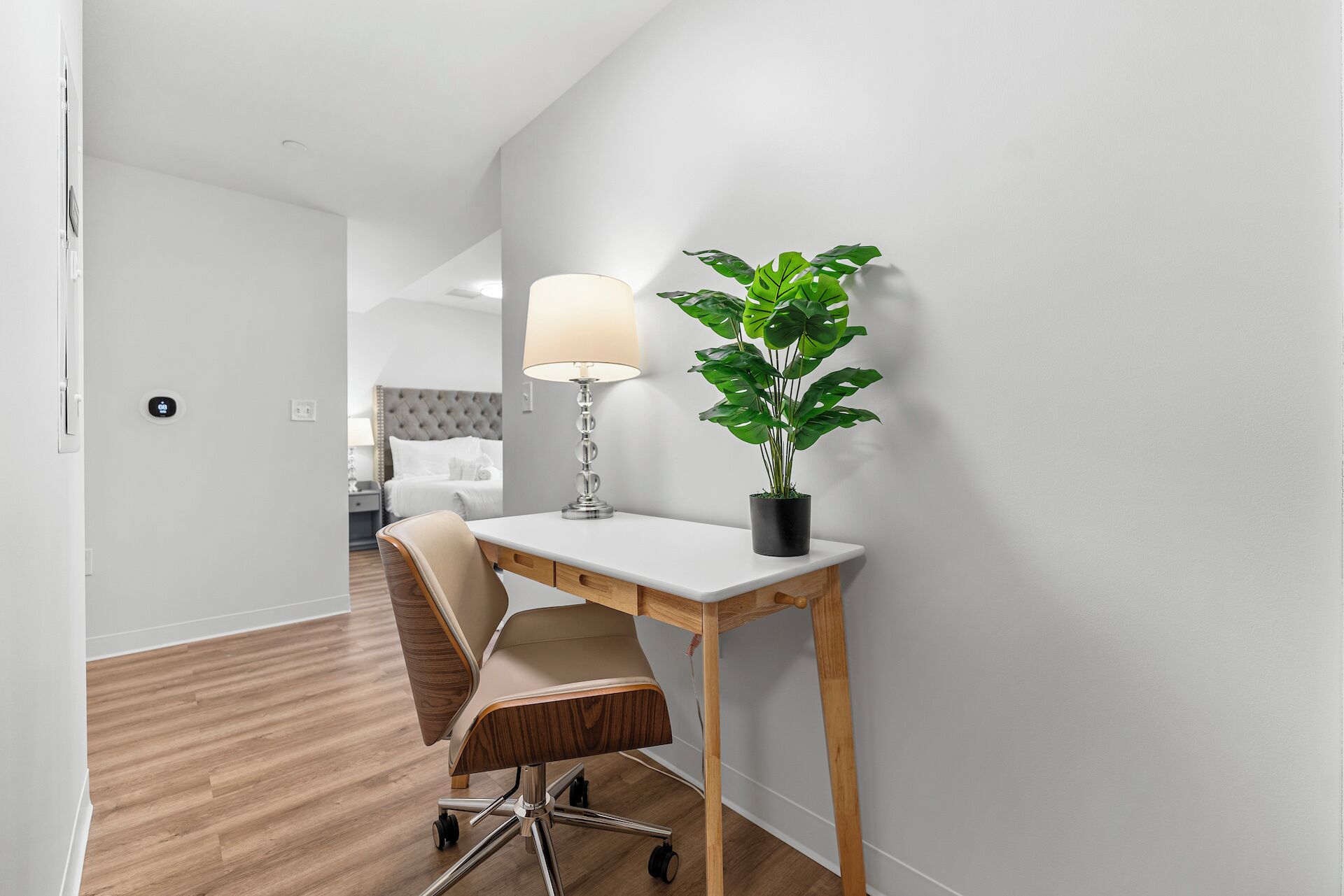 A modern hotel room with a wooden floor, a desk with a lamp, a chair, and a potted plant.