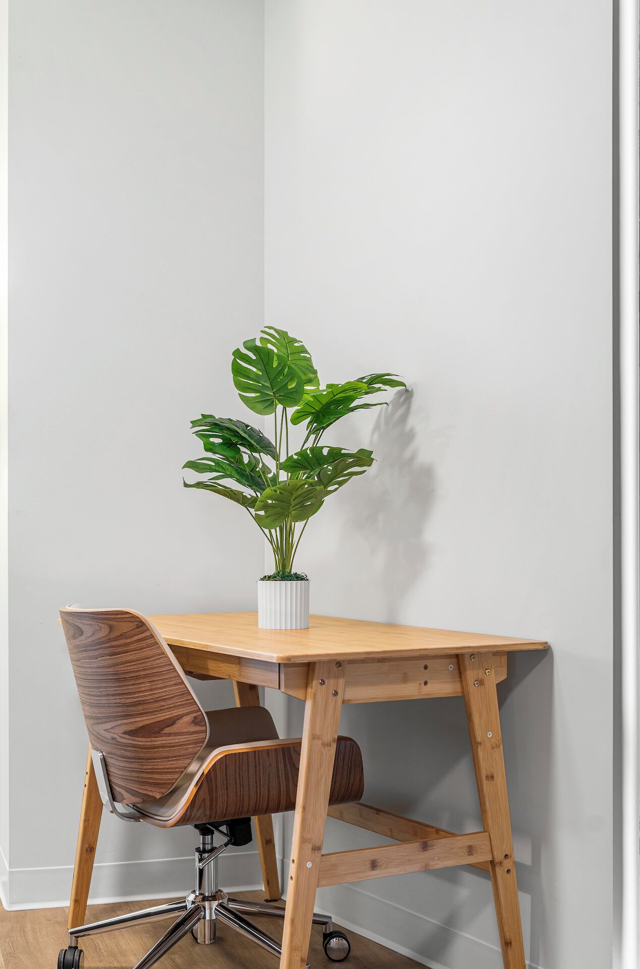 A corner desk with a potted monstera plant, a wooden chair, and a white wall.