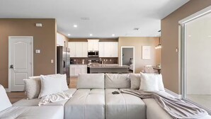 Een moderne woonkamer met een modulaire bank, een keuken op de achtergrond, en een deur die naar een andere kamer leidt.