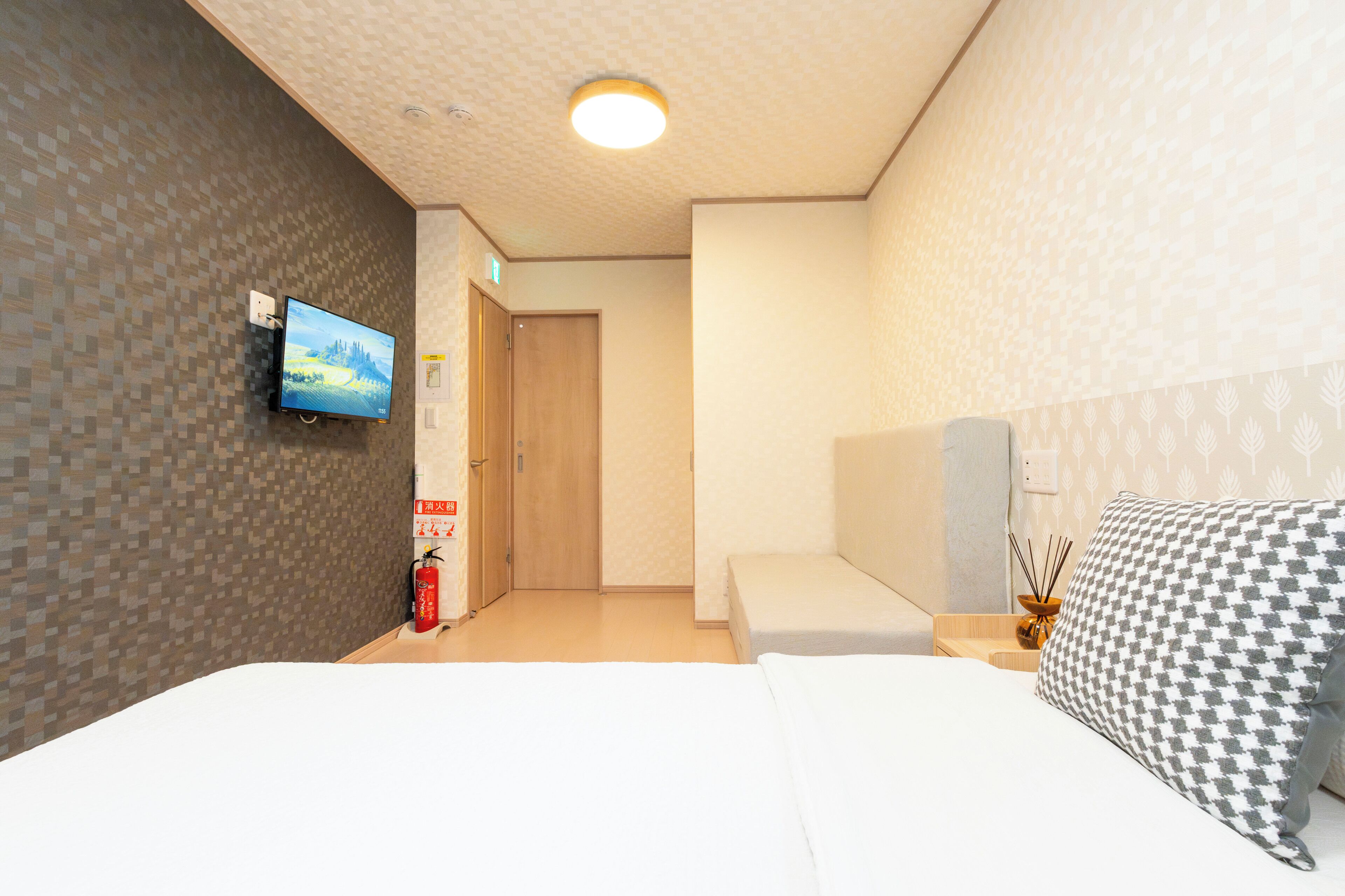 Habitación de hotel con una cama, televisor montado en la pared, mesita de noche con lámpara y una puerta que da a otra habitación.