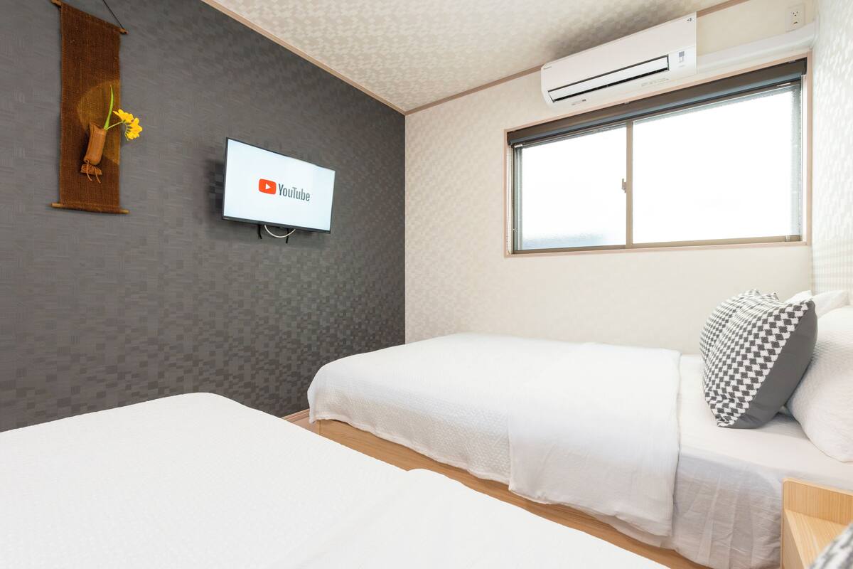 A room with two beds, a mounted TV displaying the YouTube logo, an air conditioning unit, and a window with blinds.