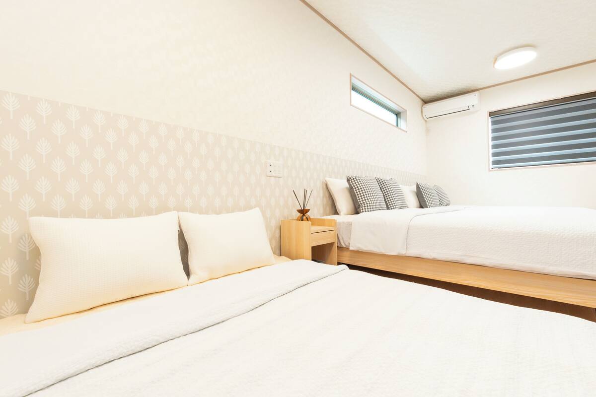 A bedroom with two beds, a small bedside table with a lamp, and a window with blinds.