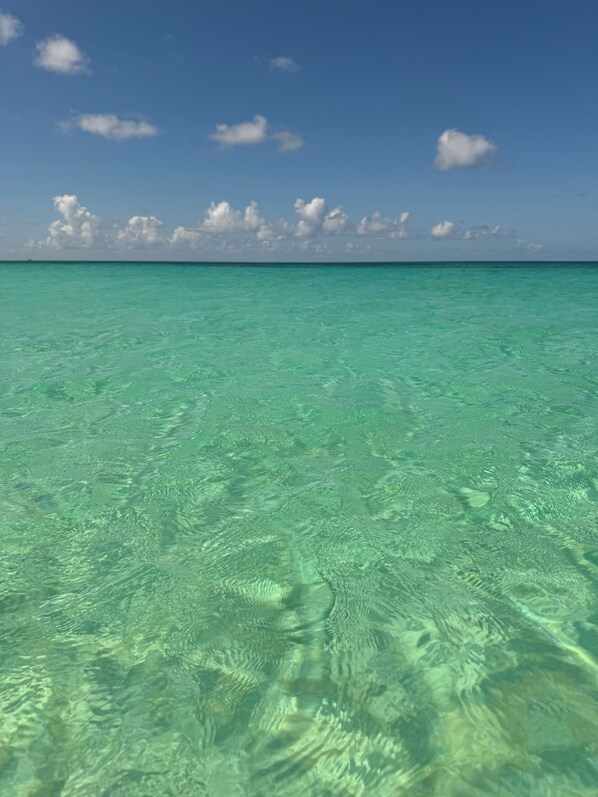 A clear view of turquoise ocean water with visible underwater patterns and a distant horizon under a blue sky with scattered clouds.
