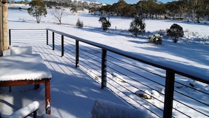 A snow-covered deck with a railing, a bench, and a view of a snowy landscape with trees and a hill in the distance.