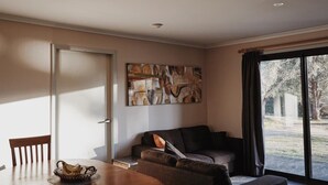 A living room with a sofa, a dining table with chairs, a painting on the wall, and a window with a view of trees.