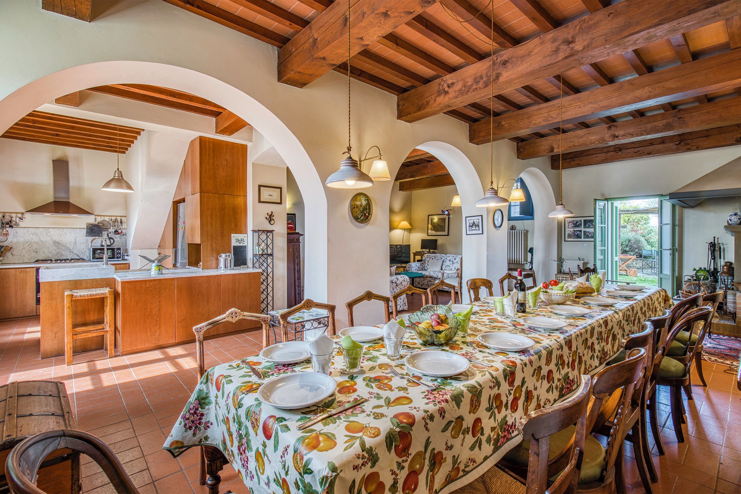 A spacious dining area with a long table set for a meal, wooden chairs, and a kitchen in the background.
