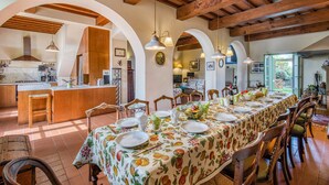 Un comedor espacioso con una larga mesa dispuesta para una comida, sillas de madera y una cocina al fondo.
