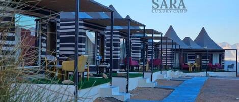 A row of modern, striped, tent-like structures with outdoor seating, set against a desert backdrop with pyramids.