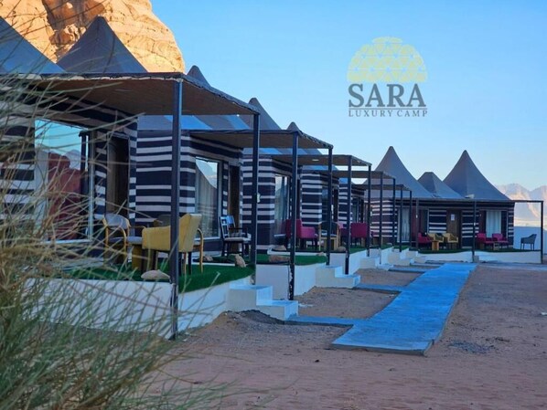 A row of modern, striped, tent-like structures with outdoor seating, set against a desert backdrop with pyramids. - Sara Luxury Camp (Wadi Rum)