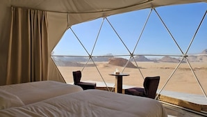 Une pièce aménagée avec une tente, comprenant un lit, deux chaises et une petite table, et offrant une vue sur un paysage désertique.