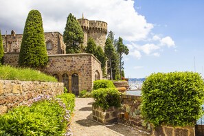 Um edifício histórico de pedra com uma torre, rodeado por jardins bem cuidados e um caminho de calçada que leva a um corpo de água.