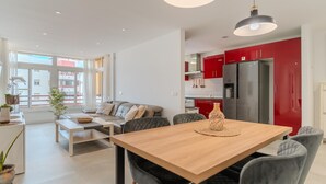 A modern living room with a sofa, coffee table, and dining table. There is a kitchen with red cabinets and a refrigerator. - Two-bedroom apartment with pool and tennis court (Costa Adeje)