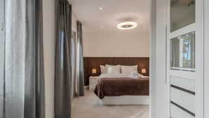 A modern hotel room with a large bed, a nightstand, a bedside table with a lamp, and a view of palm trees through a window. - Two-bedroom apartment with pool and tennis court (Costa Adeje)