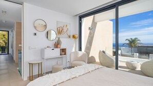 A modern bedroom with a large bed, a desk, and a chair, featuring a large window with a view of the ocean.