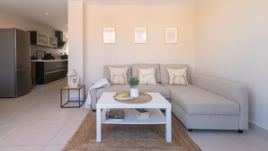 En moderne stue med en sofa, et sofabord og et lille bord. Der er et køkken i baggrunden med et køleskab og skabe.