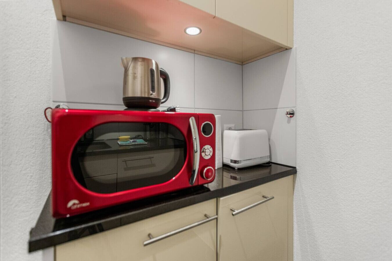Eine kleine Küchenzeile mit einer roten Mikrowelle, einem Wasserkocher und einem weißen Toaster auf einer schwarzen Arbeitsplatte.
