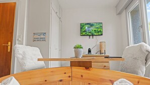 A room with a wooden dining table, two white chairs, a television mounted on the wall, a sign on the door, and a window with a view of greenery. - Crans-Montana center : Studio Ski 2 pers (Crans-Montana)