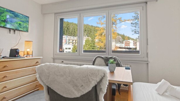 A hotel room with a large window, a wooden dresser, a chair, a small table with a plant, and a bed. - Crans-Montana center : Studio Ski 2 pers (Crans-Montana)
