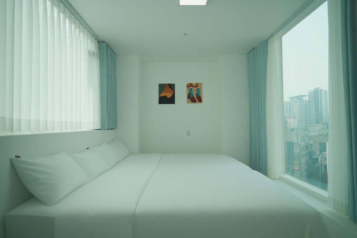 A hotel room with a large bed, two windows with curtains, and two paintings on the wall.