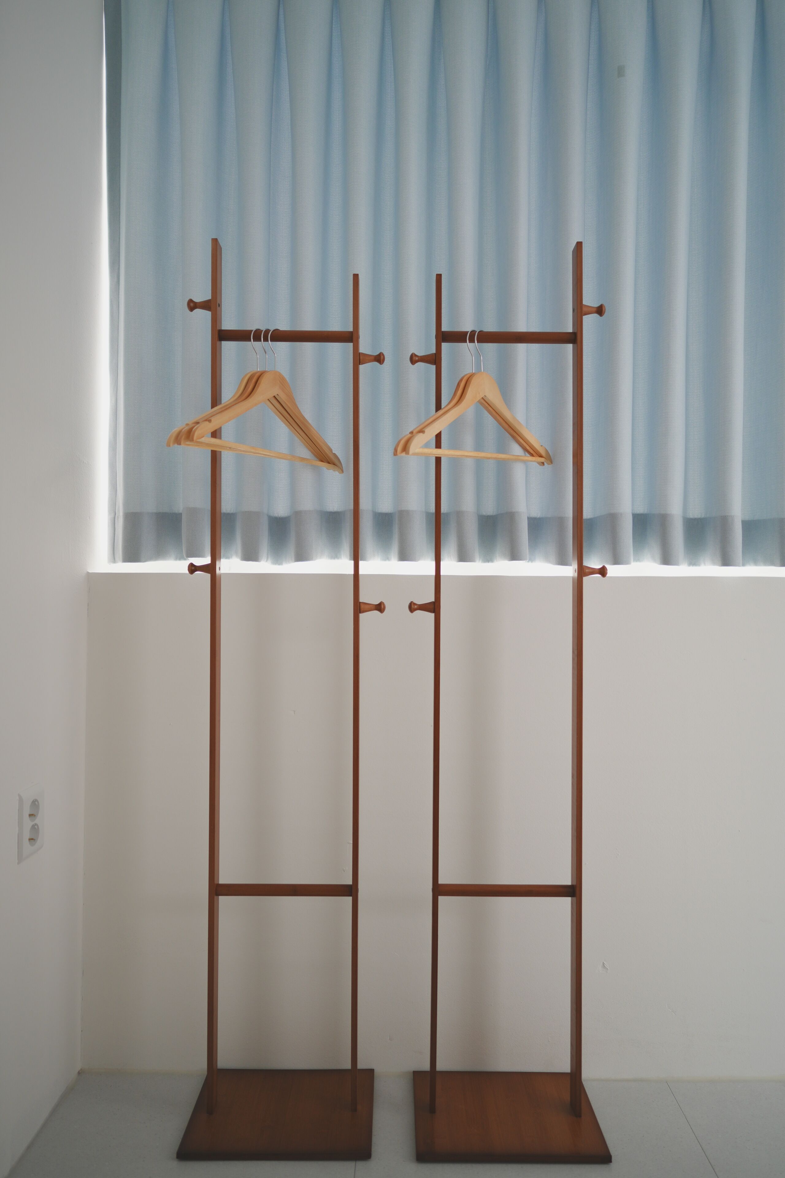 A wooden coat rack with two hangers against a window with blue curtains.