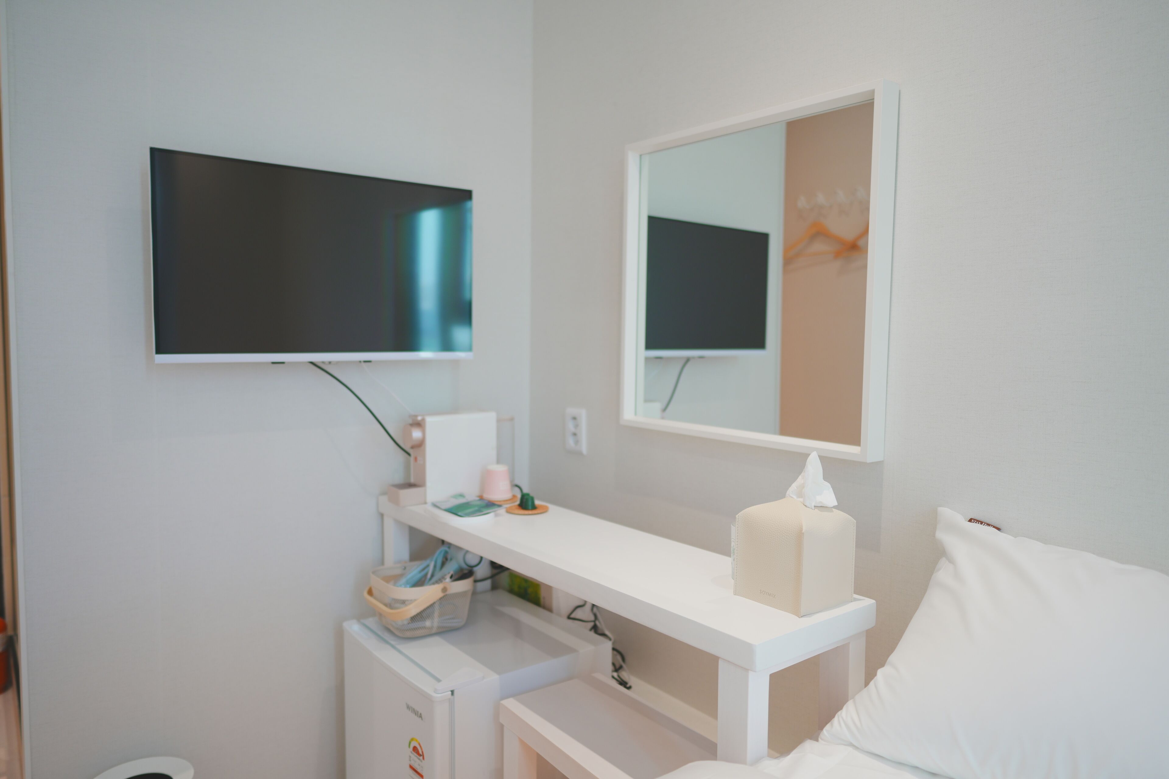 A clean, modern bathroom with a white sink, mirror, and toilet. A TV is mounted on the wall, and there is a small shelf with toiletries.