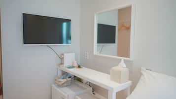 A clean, modern bathroom with a white sink, mirror, and toilet. A TV is mounted on the wall, and there is a small shelf with toiletries.